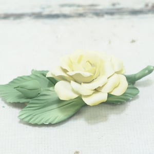 May include: A delicate porcelain rose with a pale yellow hue and green leaves. The rose is a single bloom with a closed bud on the stem.
