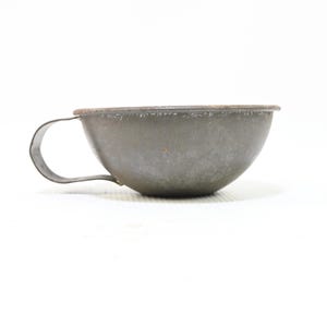 May include: A small, weathered metal cup with a rounded bowl shape and a single curved handle attached to the side. The surface of the cup displays a dull, greyish-brown patina with visible signs of oxidation and wear along the top rim.