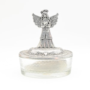 Puede incluir: Un tarro de cristal transparente con una superficie texturizada y una tapa decorativa plateada. La tapa presenta una figura de ángel con alas extendidas y un halo. El ángel sostiene un corazón. El tarro es probablemente para guardar objetos pequeños.