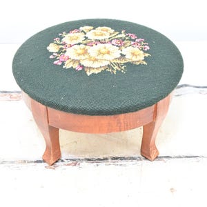 May include: A small, round footstool with a dark green fabric top featuring a floral needlepoint design in shades of yellow, pink, and white. The stool has a wooden frame and legs, showcasing a natural wood finish.