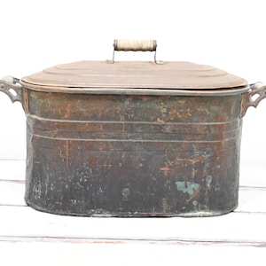 May include: An antique copper wash tub with a lid. The oval-shaped tub has a weathered, aged appearance with a patina of brown and green. It features two side handles and a wooden handle on the lid. A vintage home decor item.