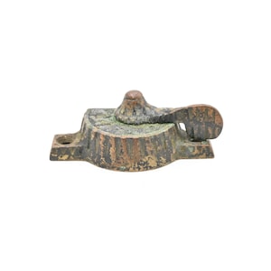 May include: A weathered bronze window latch with a lever and a small knob. The latch has a rectangular base with a hole on one side. The metal has a textured surface and a greenish patina.