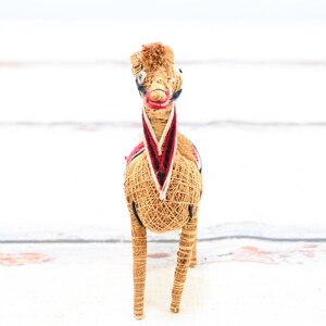 May include: A straw horse figurine with a red, white, and blue patterned saddle. The horse is standing on a white surface.
