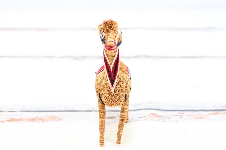 May include: A straw horse figurine with a red, white, and blue patterned saddle. The horse is standing on a white surface.