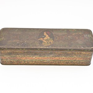 Puede incluir: Caja metálica rectangular antigua con acabado envejecido. La caja presenta un retrato ovalado central de una mujer con pañuelo rojo. La caja tiene patrones florales y geométricos decorativos en rojo, verde y dorado.