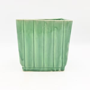 May include: A square, light green ceramic planter with vertical ribbed detailing. The planter has a slightly flared base and a smooth, glossy finish. Ideal for small plants or succulents.