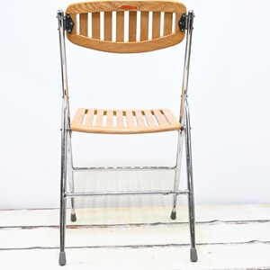 May include: A vintage folding chair with a light wood seat and backrest. The backrest is curved with vertical slats, and the seat has horizontal slats. The frame is chrome, and the chair has gray rubber feet. A classic piece of furniture.