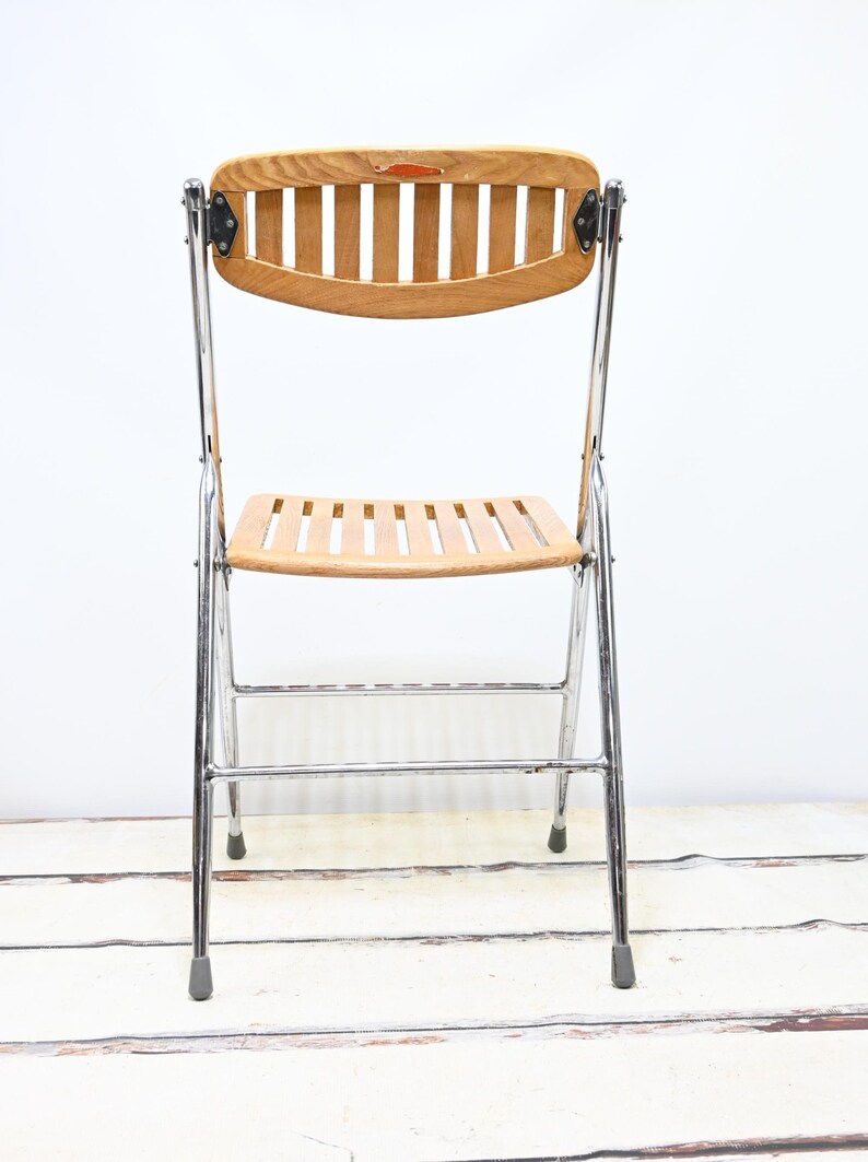 May include: A vintage folding chair with a light wood seat and backrest. The backrest is curved with vertical slats, and the seat has horizontal slats. The frame is chrome, and the chair has gray rubber feet. A classic piece of furniture.