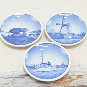 Puede incluir: Tres platos pequeños de cerámica blanca con diseños azules y blancos. Los platos presentan imágenes de un molino de viento, un barco y una formación rocosa.