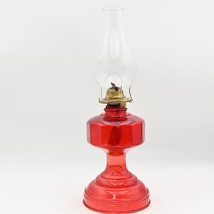 Puede incluir: Una lámpara de aceite roja vintage con una chimenea de vidrio transparente. La lámpara tiene un quemador de latón y una base facetada. La lámpara está diseñada para colocarse sobre una superficie plana. Es una pieza clásica de decoración.