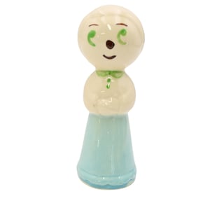 Puede incluir: Una figura de cerámica brillante de una persona estilizada con cabeza redonda, ojos pintados de verde, nariz marrón y boca pequeña marrón. La figura viste una parte superior color crema con cuello verde y una falda azul claro con borde festoneado.