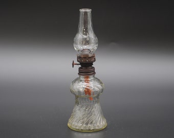 May include: A clear glass oil lamp with a twisted glass base and a metal wick holder. The lamp is on a grey surface.