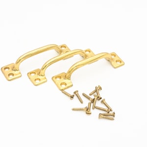 May include: Three gold-colored, arched drawer pulls with square mounting plates and screw holes are displayed. A collection of small, gold-colored screws is scattered nearby. The hardware is set against a white background.