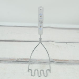 May include: A silver potato masher with a white handle. The handle has a blue and gold floral design.