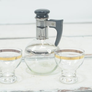 May include: A vintage glass decanter with a black plastic handle and silver accents, along with two matching glasses with gold trim.