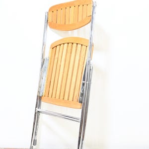 May include: A folding chair with a light wood seat and backrest, featuring vertical slats. The frame is made of shiny chrome metal. The chair is partially folded, showing its compact design. The chair is designed for indoor or outdoor use.