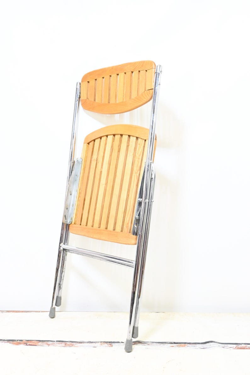 May include: A folding chair with a light wood seat and backrest, featuring vertical slats. The frame is made of shiny chrome metal. The chair is partially folded, showing its compact design. The chair is designed for indoor or outdoor use.