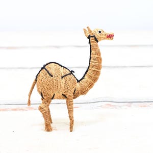 May include: A small, straw-like camel figurine with black accents and red painted lips. The camel is standing on a white surface.