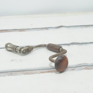 Puede incluir: Un taladro de mano vintage con mango de madera y manivela de metal. El taladro es de metal y tiene un acabado plateado. El mango es de madera oscura y tiene forma redonda.