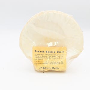 May include: A French baking shell, a round, off-white shell, is wrapped in clear plastic. A yellow label with black text reads "French Baking Shell" and describes its uses for baking and serving food. The shell is a gift from Florida.