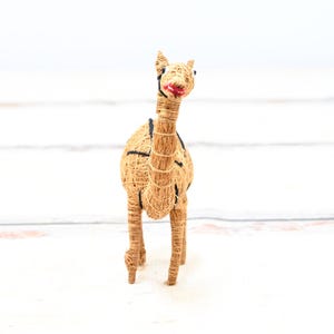 May include: A brown camel figurine made of woven straw with black eyes and a red painted mouth. The camel is standing on a white wooden surface.