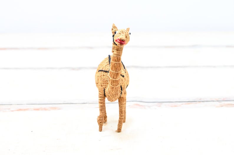 May include: A brown camel figurine made of woven straw with black eyes and a red painted mouth. The camel is standing on a white wooden surface.