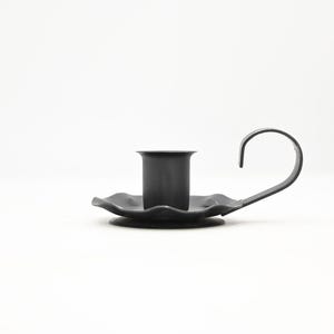 Puede incluir: Un candelabro de metal negro mate con una copa cilíndrica central para una vela, una base circular con borde festoneado y un asa curva que se extiende desde el lateral para facilitar su transporte. El diseño es sencillo y funcional.