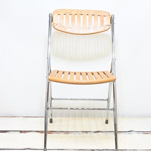May include: A vintage folding chair with a light wood seat and backrest. The chair has a chrome-colored metal frame and a slatted design on the seat and back. The chair is designed to fold for easy storage.