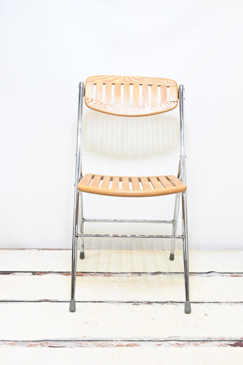 May include: A vintage folding chair with a light wood seat and backrest. The chair has a chrome-colored metal frame and a slatted design on the seat and back. The chair is designed to fold for easy storage.