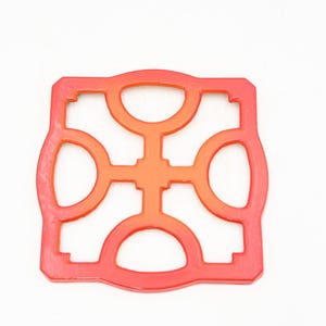 May include: A square, openwork trivet in shades of red and orange. The trivet has a repeating pattern of rounded shapes and is designed to protect surfaces from hot cookware. The trivet is made of a flexible material.