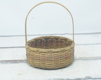 May include: A small, round, woven basket with a natural brown color and a single handle. The basket has a woven pattern with red and green accents.