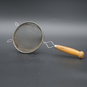 May include: A classic metal sieve with a fine mesh and a wooden handle. The handle is light brown and attached to the sieve with a metal bracket. This kitchen utensil is designed for straining liquids or sifting ingredients.