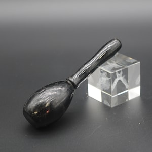 Puede incluir: Un mazo de madera negro con una cabeza redonda y un mango largo y delgado. El mazo está sobre una superficie negra junto a un cubo de vidrio transparente.