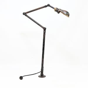 May include: A dark metal articulated desk lamp with a long, adjustable arm and a circular base. The lamp features a weathered, industrial finish with visible joints and a small, rounded shade at the end of the arm, connected by a black cord.