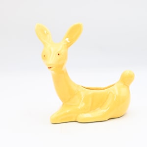 May include: A yellow ceramic planter in the shape of a deer. The deer has long ears, a small red nose, and a curved body with a small tail. The planter is designed to hold small plants or flowers.