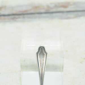 May include: Close-up of the top of a silver spoon with a decorative, ornate design.
