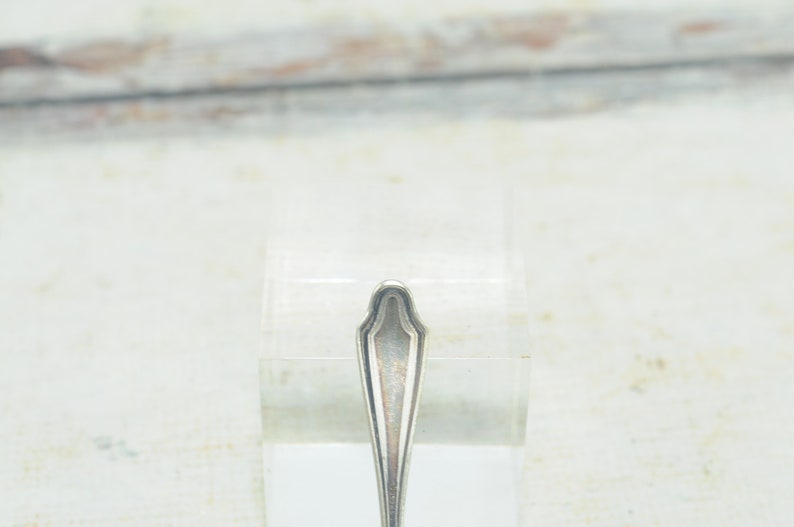 May include: Close-up of the top of a silver spoon with a decorative, ornate design.
