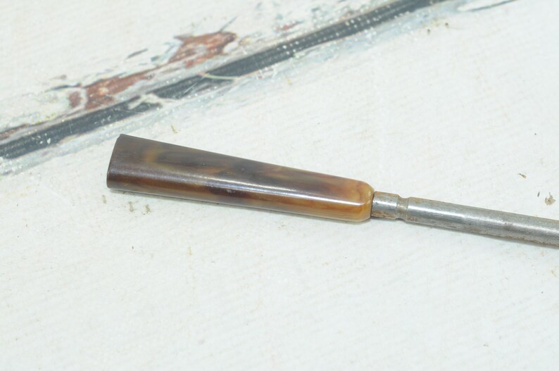 Op de afbeelding: Een metalen gereedschap met een lange, dunne schacht en een bruin en beige handvat.
