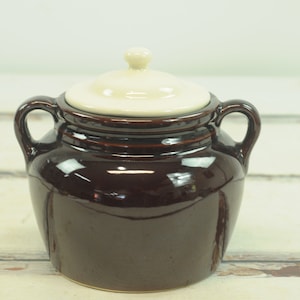 Puede incluir: Una olla de frijoles de cerámica marrón con una tapa blanca y dos asas. La olla está esmaltada y tiene un acabado brillante.