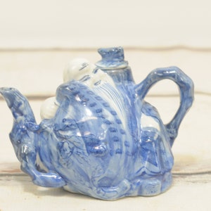 Könnte beinhalten: Blaue und weiße Keramik-Teekanne mit einem Design einer chinesischen Gottheit. Die Teekanne hat einen Ausguss und einen Griff.