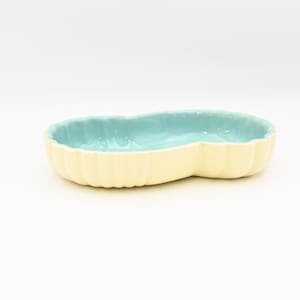 May include: A light yellow ceramic dish with a light blue interior. The dish has a unique, elongated shape with a scalloped edge. The dish is on a white background.