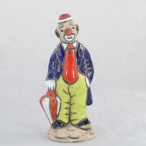 May include: A ceramic clown figurine with a red hat, blue jacket, red tie, and yellow pants. The clown is holding a red and white umbrella.