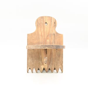 Puede incluir: Un especiero rústico de madera para montar en la pared, con un acabado natural. El especiero presenta una parte superior decorativa, un estante y un borde inferior con dientes puntiagudos. La veta de la madera es visible.