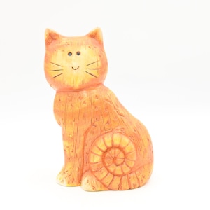 Op de afbeelding: Oranje keramieken kattenbeeldje met een lachend gezicht en zwarte snorharen. De kat zit in een zittende positie met een spiraalvormig ontwerp aan de zijkant. Het beeldje heeft een getextureerd oppervlak en staat op een witte achtergrond.
