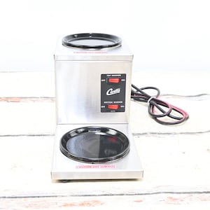 May include: A stainless steel Curtis coffee warmer with two black warming plates. It has red on/off switches for the top and bottom warmers. A black and red power cord is attached.