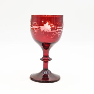 May include: A ruby red wine glass with a decorative etched floral design. The glass has a rounded bowl, a slender stem, and a circular base. The etched design features a flower and vines.