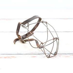 May include: A vintage dog muzzle made of metal wire and leather straps. The muzzle is designed to fit over the dog's snout and has a wire cage to prevent biting.