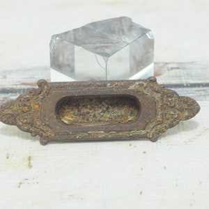 Puede incluir: Un tirador de puerta de metal oxidado con detalles ornamentados. El tirador tiene una abertura rectangular y está diseñado para ser utilizado como manija para una puerta o un cajón.