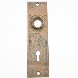 Könnte beinhalten: Eine rechteckige Türschildplatte aus Bronze mit verwitterter Patina, einer zentralen runden Öffnung für den Türknauf mit geriffeltem Kragen und einem Schlüsselloch darunter. Die Oberfläche ist mit geometrischen Linien und Pflanzenmotiven verziert.