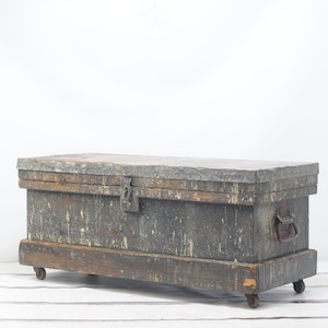May include: A vintage wooden storage chest with a distressed gray paint finish. The chest has a hinged lid, a metal latch, and a metal handle. It sits on four small wooden wheels.